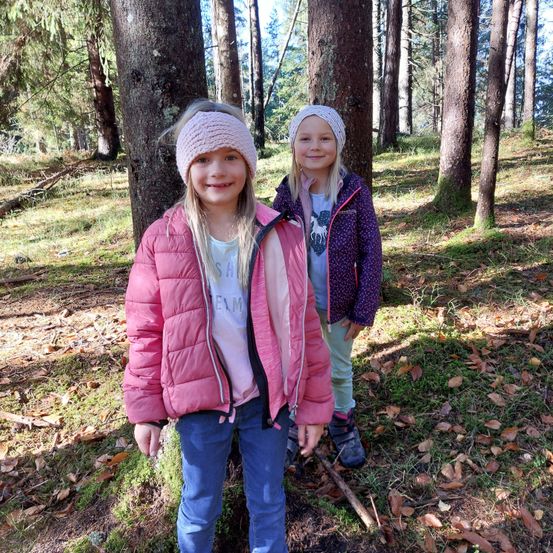 Zwei junge Mädchen stehen lächelnd in einem Wald. Eine trägt eine rosa Jacke, die andere eine lila Jacke mit weißer Stirnband. Sie sind von hohen Bäumen und gefallenen Blättern umgeben.