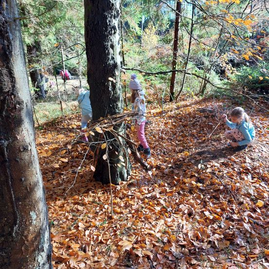 Drei Kinder sind in einem Wald mit gefallenen Blättern. Sie spielen mit Stöcken und Zweigen, und ein Kind sitzt auf dem Boden.