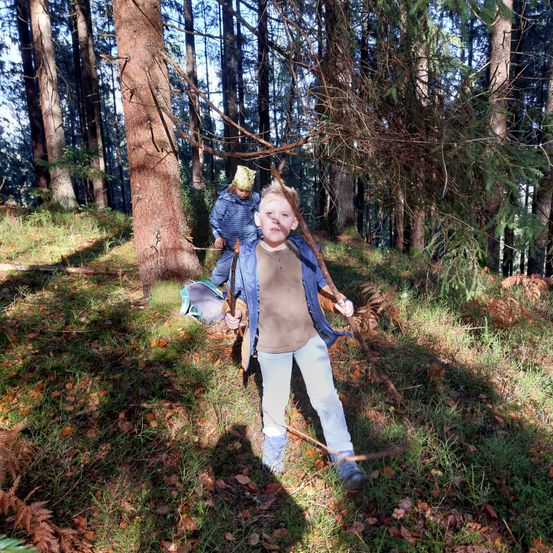 Zwei Kinder gehen mit Stöcken bewaffnet durch einen Wald und tragen Jacken. Ein Kind hat einen gelben Hut, und sie sind von hohen Bäumen umgeben.