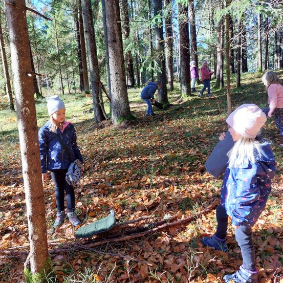 Eine Gruppe von Kindern erkundet ein bewaldetes Gebiet. Sie tragen Jacken und Mützen, wobei ein Kind einen Stock hält. Der Boden ist mit gefallenen Blättern bedeckt.