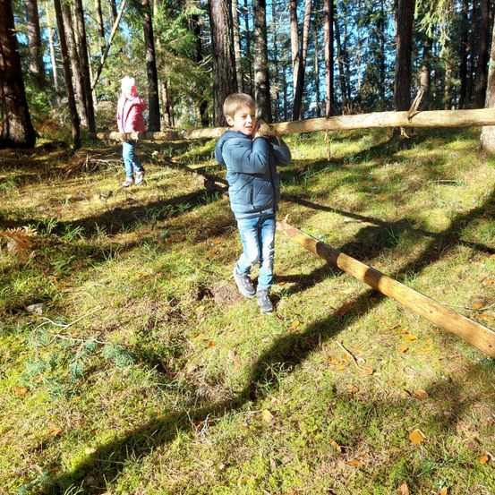 Zwei Kinder spielen im Wald, eines hält einen Baumstamm, während das andere hinter ihm steht. Sie sind von hohen Bäumen umgeben.