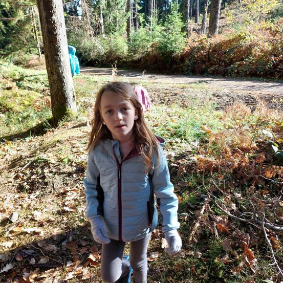 Ein junges Mädchen steht in einem Wald, umgeben von Laub, trägt eine hellblaue Jacke mit rotem Reißverschluss und weiße Handschuhe.