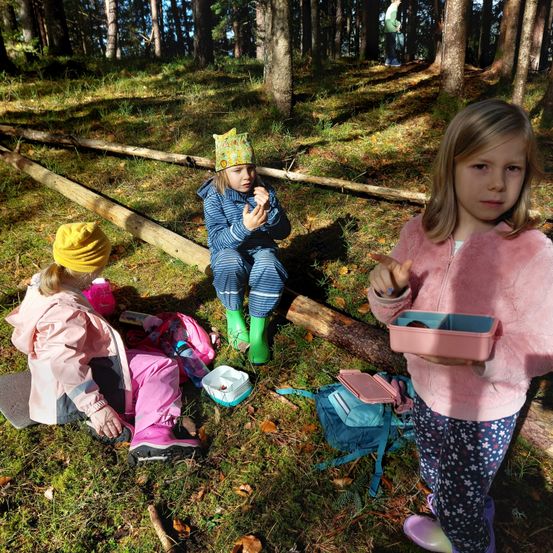 Drei kleine Mädchen sitzen in einem Wald, eines mit einer rosa Brotdose. Sie sind in Regenkleidung und Stiefeln gekleidet. Ein anderes Kind steht im Hintergrund.