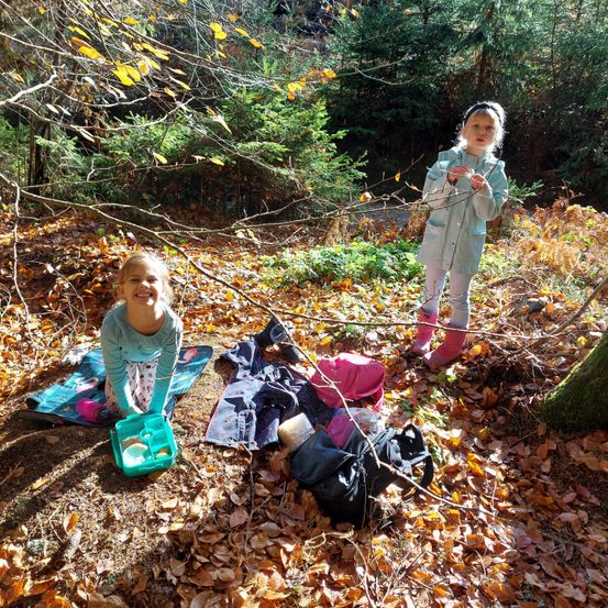 Zwei junge Mädchen spielen im Wald. Eine sitzt auf einer Decke und hält einen Behälter, während die andere steht und einen Zweig hält. Sie sind von gefallenen Blättern und Bäumen umgeben.