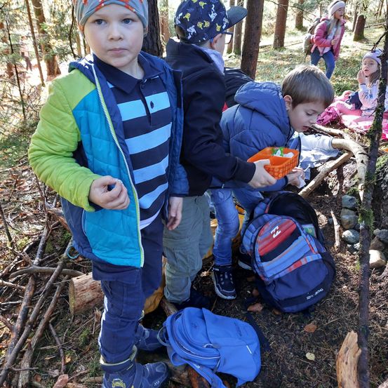 Eine Gruppe von Kindern ist in einem Wald, sie schauen sich etwas auf dem Boden an, vielleicht eine Schatzsuche. Sie tragen Winterkleidung und haben Rucksäcke dabei.