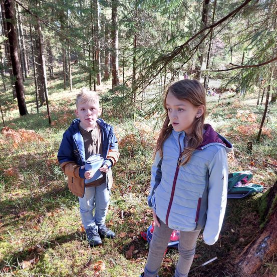 Zwei Kinder stehen in einem Wald, ein Junge hält einen blauen Behälter und ein Mädchen trägt eine Jacke mit Kapuze.
