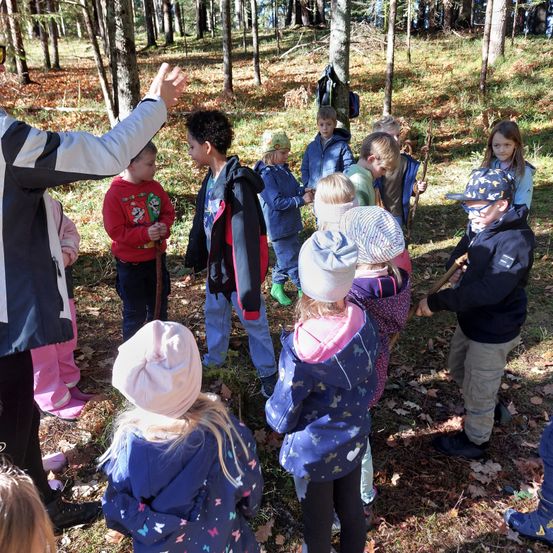Eine Gruppe von Kindern und einem Lehrer ist in einem Wald. Die Kinder halten Stöcke und tragen Jacken und Mützen. Der Lehrer trägt eine weiße Jacke.