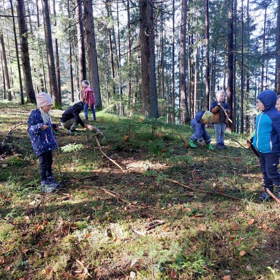 Eine Gruppe von Kindern erkundet einen Wald. Sie halten Stöcke und durchsuchen den Boden. Einige tragen Mützen und Stiefel. Der Wald ist von hohen Bäumen umgeben.