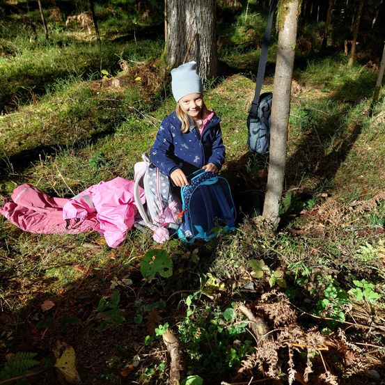 Ein kleines Mädchen mit Rucksack und Mütze sitzt in einer Waldlichtung neben einem Baumstamm, mit ihren Kleidern auf dem Boden ausgebreitet.