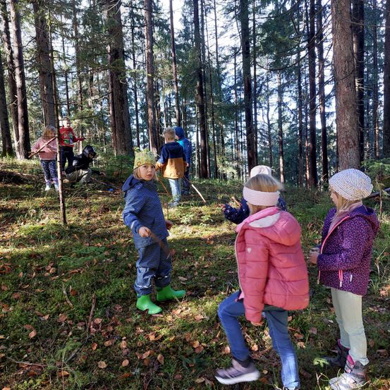 Kinder sind in einem Wald, einige halten Stöcke. Ein Mädchen in grünen Stiefeln sticht hervor. Andere tragen Mützen und dicke Jacken. Sie befinden sich auf einem Rasen mit gefallenen Blättern.