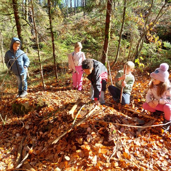 Fünf Kinder befinden sich in einem Wald, einige sammel Zweige, und eines steht auf einem Baumstumpf. Sie tragen Winterkleidung, und der Boden ist mit Laub bedeckt.