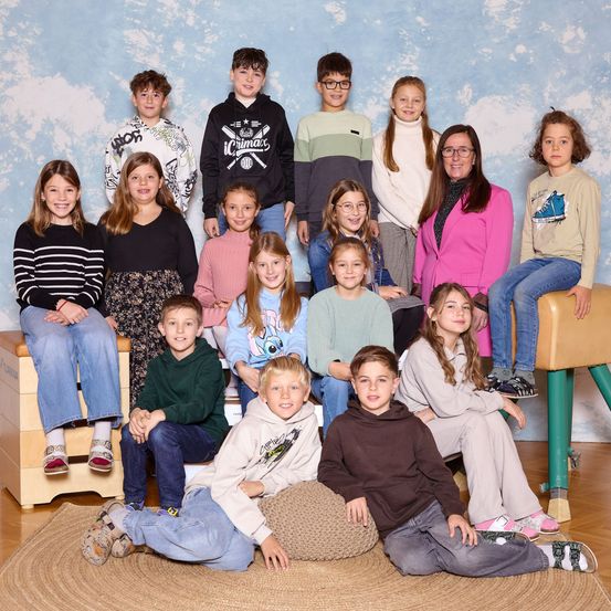 Eine Gruppe junger Schüler, Jungen und Mädchen, posiert für ein Schulfoto. Einige sitzen auf einer Bank, andere knien auf einem Teppich. Sie alle lächeln in die Kamera.