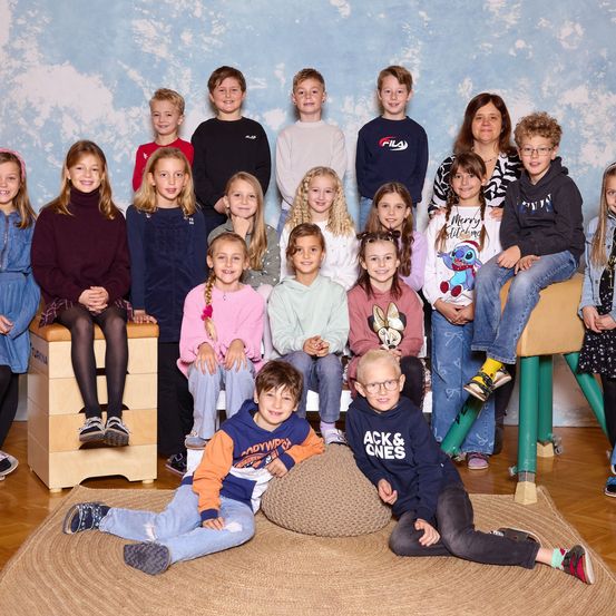 Eine Gruppe von Kindern und zwei Erwachsenen posiert für ein Foto. Sie sitzen und stehen in Reihen. Ein Kind sitzt auf einem Plüschhocker. Die Wand hinter ihnen hat ein Himmelsdesign.