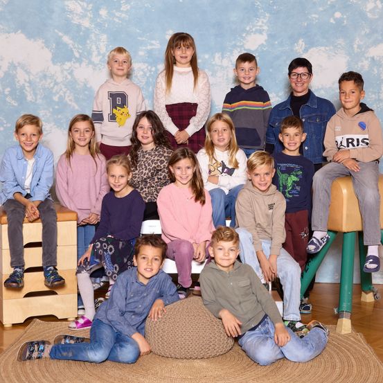 Eine Gruppe von Kindern und zwei Erwachsenen posiert für ein Foto vor einem blauen Hintergrund. Die Kinder sitzen und stehen, tragen Freizeitkleidung. Einige haben Turnschuhe an.
