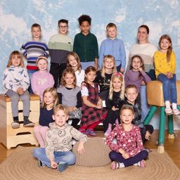 Eine Gruppe von Kindern und einer Frau posiert für ein Foto in einem Klassenzimmer. Sie sitzen auf verschiedenen Möbeln, einschließlich einer Bank und einem Hocker. Die Frau steht hinter ihnen und lächelt. Die Kinder tragen verschiedene Kleidungsstücke wie Kleider, Hemden und Hosen. Sie alle schauen in dieselbe Richtung.