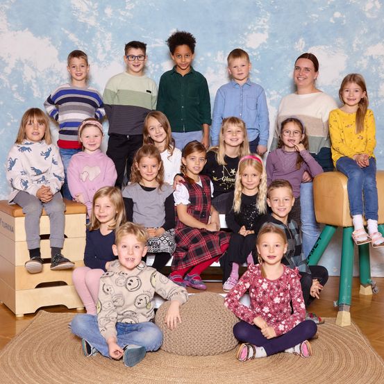 Eine Gruppe von Kindern und einer Frau posiert für ein Foto in einem Klassenzimmer. Sie sitzen auf verschiedenen Möbeln, einschließlich einer Bank und einem Hocker. Die Frau steht hinter ihnen und lächelt. Die Kinder tragen verschiedene Kleidungsstücke wie Kleider, Hemden und Hosen. Sie alle schauen in dieselbe Richtung.