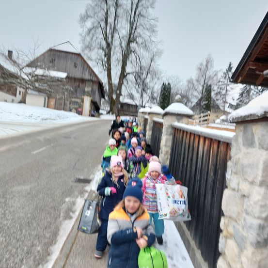 Eine Gruppe von Kindern geht mit Taschen auf einem verschneiten Straßenweg. Dahinter befindet sich ein Holzzaun und ein Gebäude mit einem schneebedeckten Dach.