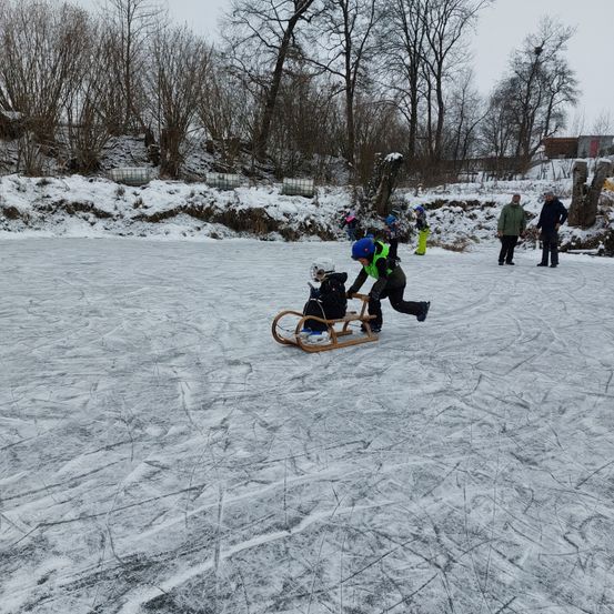 Eine Gruppe von Menschen befindet sich auf einem zugefrorenen See. Eine Person schiebt einen hölzernen Schlitten, auf dem zwei andere sitzen. Bäume und Schnee umgeben das Gebiet.