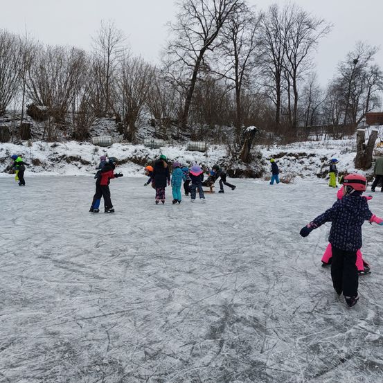Mehrere Kinder spielen Schlittschuh auf einem gefrorenen Teich in einer verschneiten Landschaft mit Bäumen und einem Hügel im Hintergrund.
