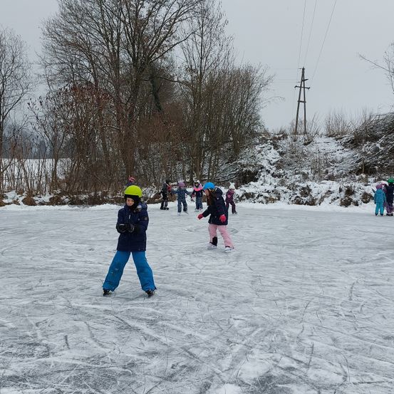 Eine Gruppe von Kindern skat auf einem zugefrorenen See, mit Helmen und Handschuhen. Sie sind von Bäumen und Schnee umgeben.