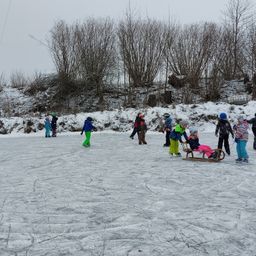 Mehrere Kinder rutschen auf einer Freiluft-Eisbahn, die von Schnee umgeben ist. Einige tragen Helme. Ein junges Mädchen fährt auf einem Schlitten, der von einem Erwachsenen gezogen wird.