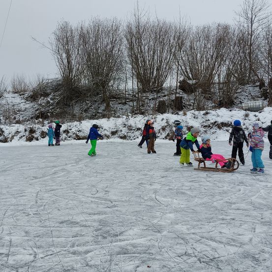 Mehrere Kinder rutschen auf einer Freiluft-Eisbahn, die von Schnee umgeben ist. Einige tragen Helme. Ein junges Mädchen fährt auf einem Schlitten, der von einem Erwachsenen gezogen wird.