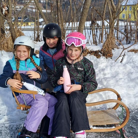 Drei junge Mädchen in Winterkleidung sitzen auf einem Schlitten in einem verschneiten Bereich und lächeln für die Kamera. Eine hält eine rosa Wasserflasche, eine andere eine weiße. Hinter ihnen stehen Bäume und ein gelbes Haus.