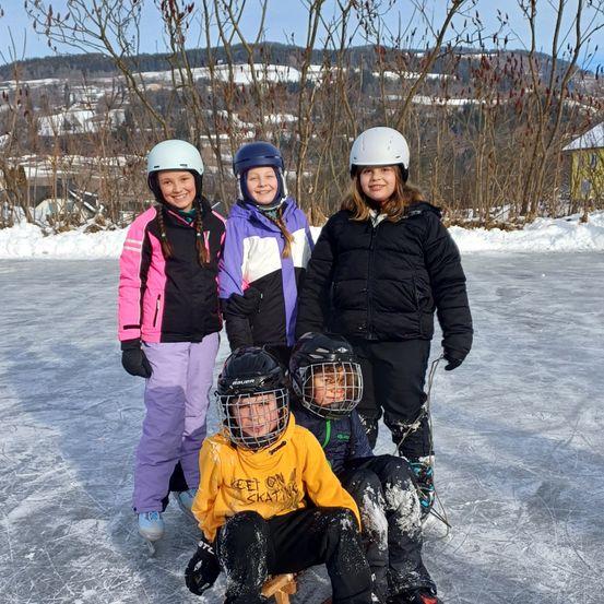Fünf Kinder, vier mit Helmen, posieren für ein Foto auf einer verschneiten Eisbahn mit einem verschneiten Berg im Hintergrund.