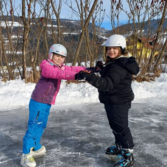 Zwei junge Mädchen in Winterkleidung rutschen auf einem zugefrorenen See. Sie lächeln und halten Händchen. Im Hintergrund stehen laublose Bäume und ein gelbes Gebäude.