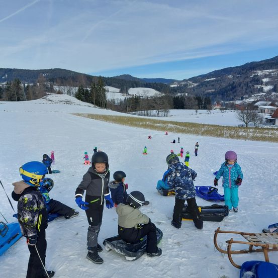 Mehrere Kinder spielen im Schnee. Einige fahren mit Schlitten, während andere herumstehen. Die schneebedeckten Berge sind im Hintergrund.