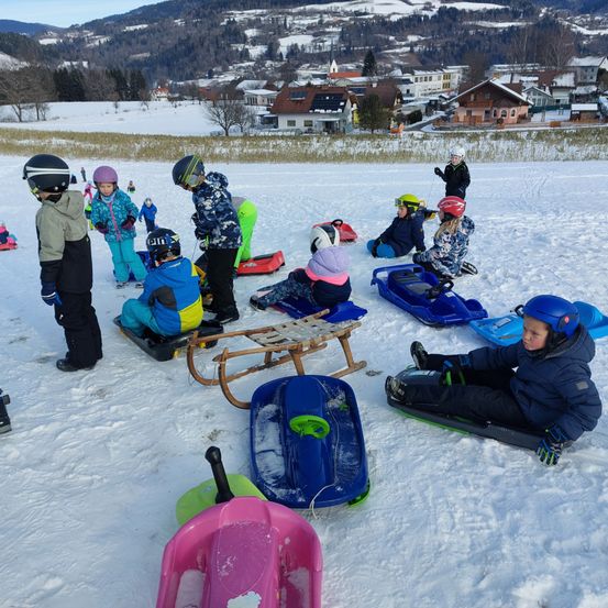 Eine Gruppe von Kindern spielt im Schnee und sitzt auf Schlitten. Einige tragen Helme und sind in Winterkleidung. Dahinter liegt eine verschneite Landschaft mit einem Haus und Bergen.