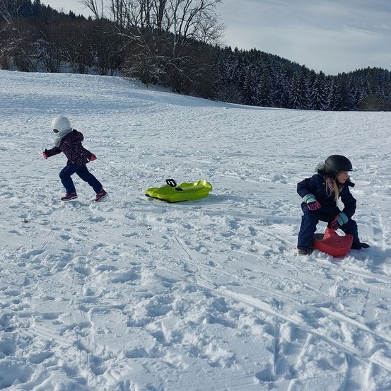 Zwei Kinder haben Spaß im Schnee, eines zieht einen grünen Schlitten und das andere sitzt auf einem roten. Sie tragen beide Helme und Handschuhe.