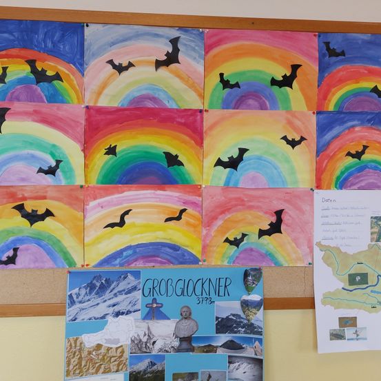 Ein Bulletinbrett zeigt ein farbenfrohes Kunstwerk mit Regenbögen und Fledermäusen. Darunter befindet sich ein Poster über den Großglockner, einen Berggipfel.