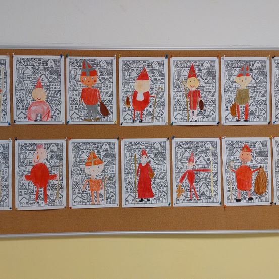 Ein Korkbrett zeigt mehrere Kinderzeichnungen, jeweils mit einer Figur in roter Kleidung, die vor Gebäudehintergrund steht. Die Figuren scheinen Teil eines Zuges oder einer Prozession zu sein.