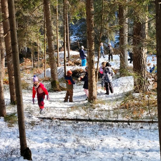Eine Gruppe von Kindern in Winterkleidung geht im verschneiten Wald. Der Boden und einige Bäume sind mit Schnee bedeckt.