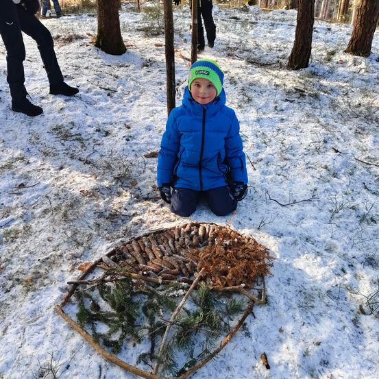 Ein Kind in einer blauen Winterjacke kniet auf schneebedecktem Boden, umgeben von einem selbstgemachten Kunstwerk aus Kiefernzapfen und Zweigen. Im Hintergrund gehen Menschen auf dem schneebedeckten Pfad.