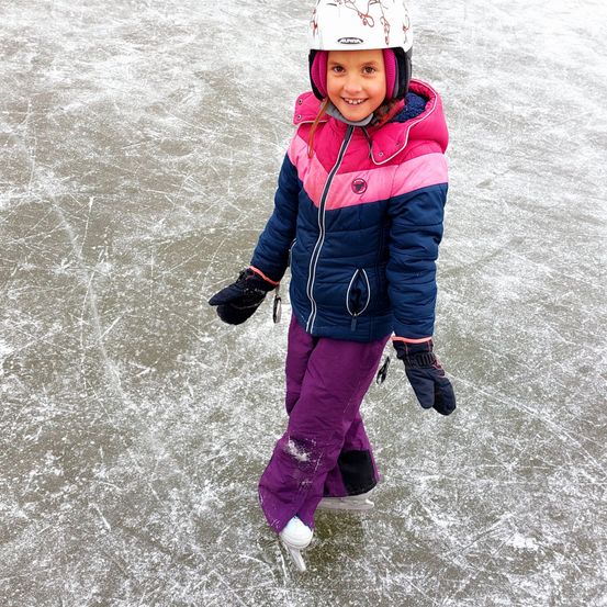 Ein junges Mädchen schreitet auf einer Eisbahn, trägt einen Helm, eine bunte Jacke, Handschuhe und Schlittschuhe. Sie lächelt in die Kamera und steht auf glattem Eis.