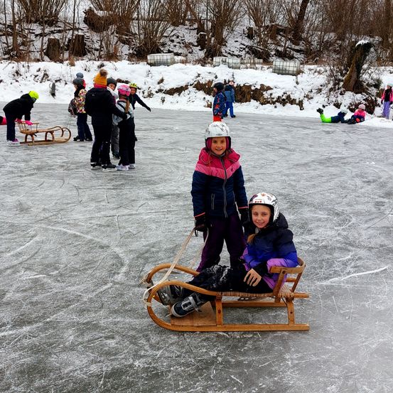 Mehrere Kinder in Winterkleidung sind auf einer Eisbahn. Zwei von ihnen fahren auf einem hölzernen Schlitten. Andere Kinder stehen und sitzen im Schnee.