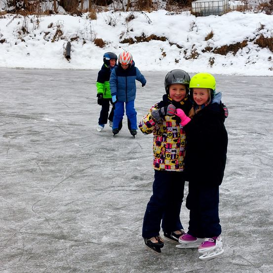 Drei Kinder sind Schlittschuhlaufen auf einem zugefrorenen See. Zwei Mädchen vorne tragen bunte Jacken und Helme. Ein Junge hinter ihnen skatet mit seinen Freunden.