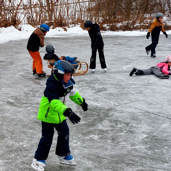 Eine Gruppe von Kindern Schlittschuh laufen auf einem gefrorenen See. Einer sitzt auf einem Schlitten, ein anderer liegt auf dem Boden, und einer hält einen Schlitten. Bäume und Schnee sind im Hintergrund.