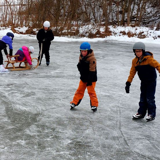 Eine Gruppe von Kindern ist auf einem gefrorenen See. Einige eislaufen, andere sitzen auf einem Schlitten. Sie tragen alle Helme und Handschuhe.