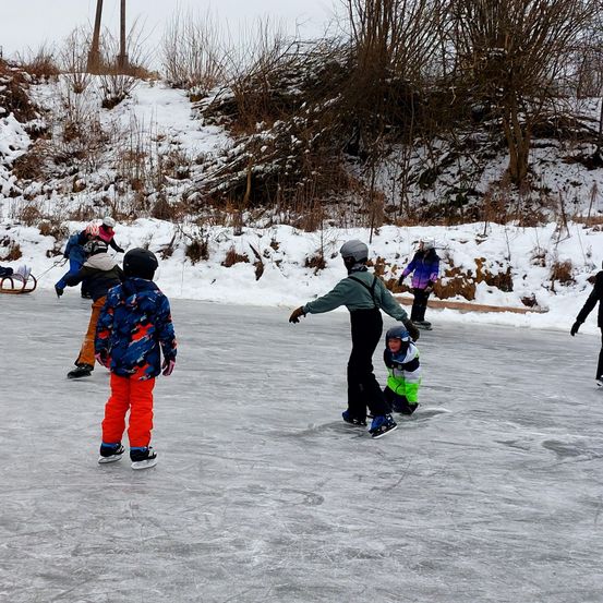 Eine Gruppe von Kindern und Erwachsenen eislaufen auf einem zugefrorenen See. Sie tragen Winterkleidung und Helme. Einer von ihnen zieht einen Schlitten.
