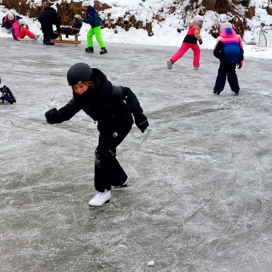 Mehrere Kinder sind auf einer eisigen Oberfläche, wahrscheinlich einer Eisbahn, einige tragen Helme. Ein Kind trägt einen schwarzen Helm und scheint zu gehen.