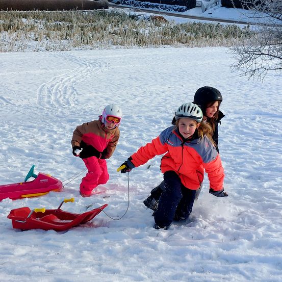 Drei Kinder in Winterkleidung und Helmen spielen im Schnee. Eines zieht einen roten Schlitten, während die anderen in der Nähe stehen.