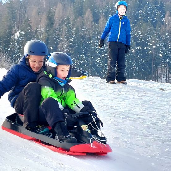 Drei Kinder in Winterkleidung fahren mit einem Schlitten einen verschneiten Hügel hinunter. Zwei sitzen auf dem Schlitten, während das dritte im Hintergrund steht.