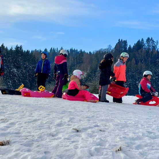 Eine Gruppe von Kindern in Winterkleidung spielt auf Schlitten auf einem verschneiten Hügel mit Kiefern im Hintergrund.