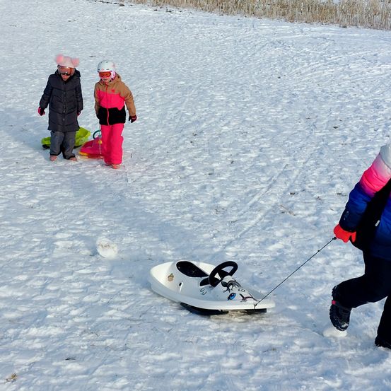 Drei Kinder in Winterkleidung ziehen einen Schlitten im Schnee. Ein Kind trägt einen schwarzen Mantel und einen rosa Hut. Ein anderes trägt eine pinke und braune Jacke. Das dritte trägt einen blauen und rosa Mantel.