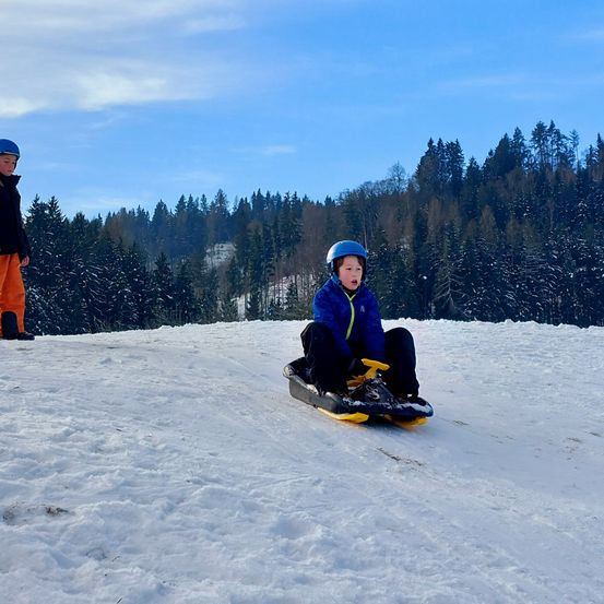 Zwei Kinder sind auf einem verschneiten Hügel. Eines sitzt auf einem Schlitten, trägt eine blaue Jacke und einen Helm, während das andere steht, gekleidet in eine schwarze Jacke und orange Hosen. Dahinter ist die Landschaft schneebedeckt, mit Kiefern im Hintergrund.