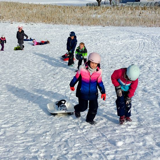 Eine Gruppe von Kindern spielt im Schnee. Einige rodeln, andere liegen. Ein Kind zieht einen Schlitten.