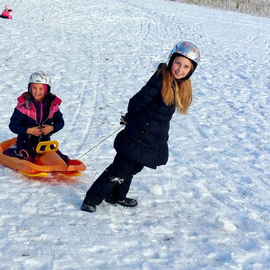 Zwei Mädchen spielen im Schnee. Eine sitzt auf einem Schlitten und die andere zieht sie. Beide tragen Winterkleidung und Helme. Das Mädchen auf dem Schlitten lächelt und hält den Schlitten mit den Händen fest.